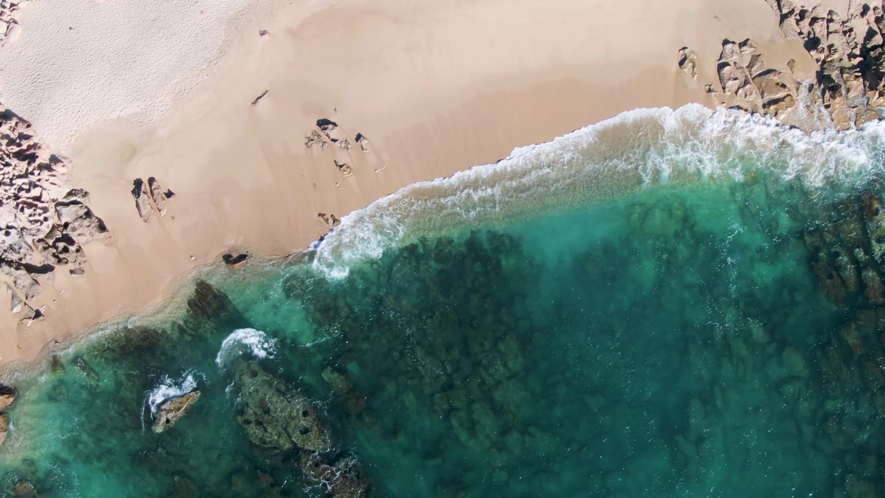 Overhead view of Buzzards Beach in Baja California Sur with turquoise water meeting pale sand, white foam at the shore, dark rocks, and small people visible on the beach.