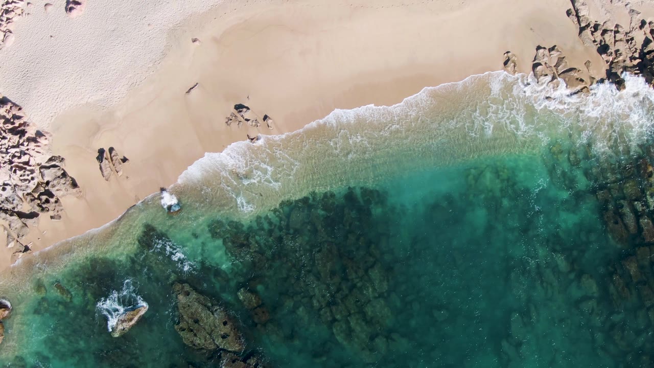 At 0:02, turquoise water fills the lower frame beneath a pale sandy beach, with white wash near the rocks and small people visible near the upper beach area at Buzzards Beach.