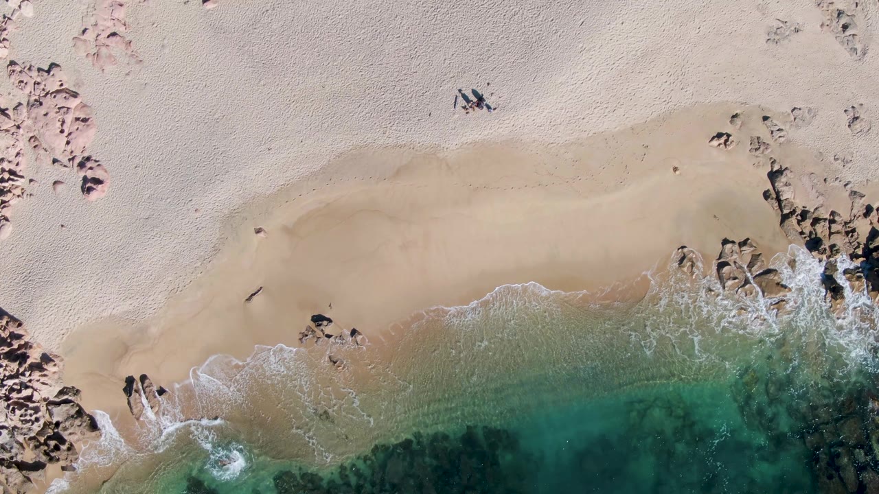 Overhead view of Buzzards Beach in Baja California Sur with pale sand, scattered rock outcrops, turquoise water at the lower edge, and a small person walking near the upper left area.