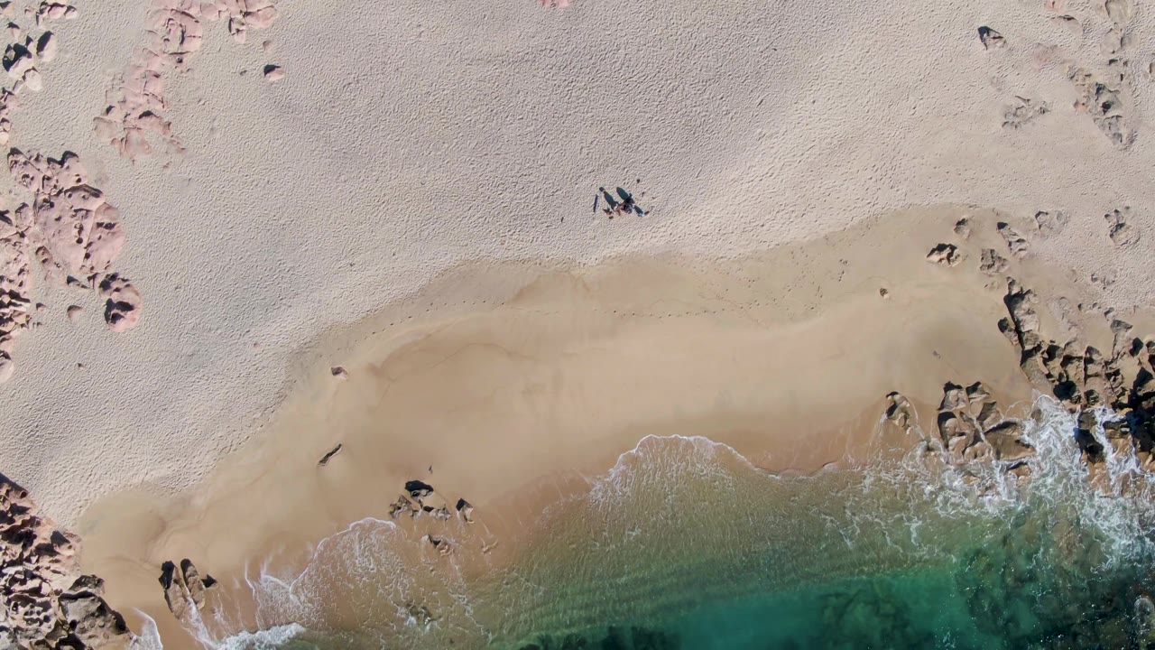 Aerial beach view with one walker, curved shoreline, rocks, and shallow water.