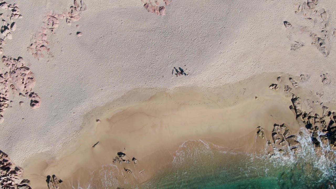 Top-down view of Buzzards Beach with broad sand flats, pink-toned rock patches near the top, a tiny person on the beach, and surf breaking around rocks at the lower right.