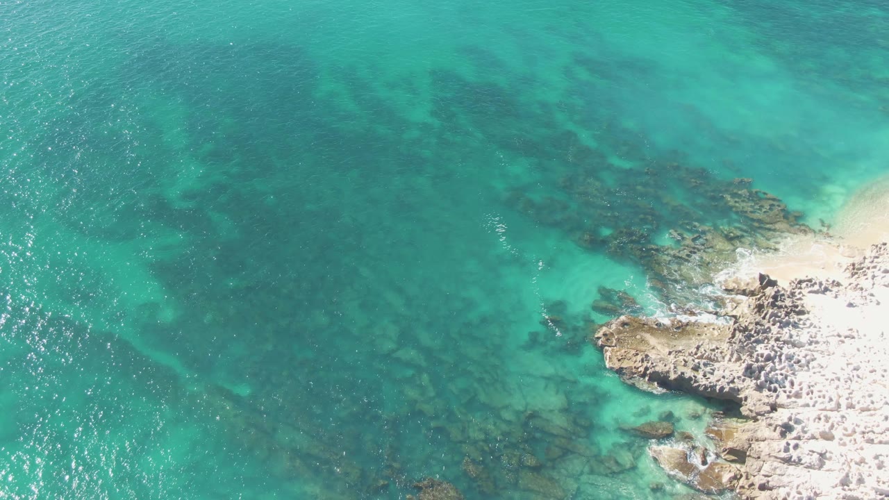 High overhead view of Buzzards Beach turquoise water with submerged rocks and a bright pale rocky shoreline entering at the lower right, with no visible people.