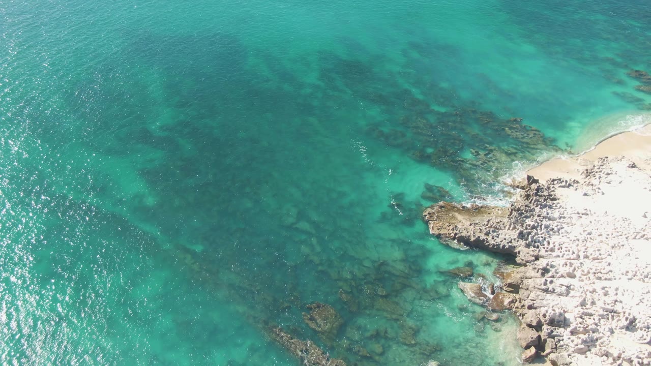Aerial view over Buzzards Beach with bright turquoise shallows, darker submerged rock formations, and a white rocky coast crossing the lower right, with no visible people.