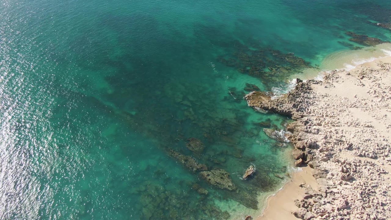Aerial frame over Buzzards Beach in Baja California Sur showing turquoise water, submerged rocks, pale sand, rocky shoreline, sun sparkle on the sea, and no visible people.