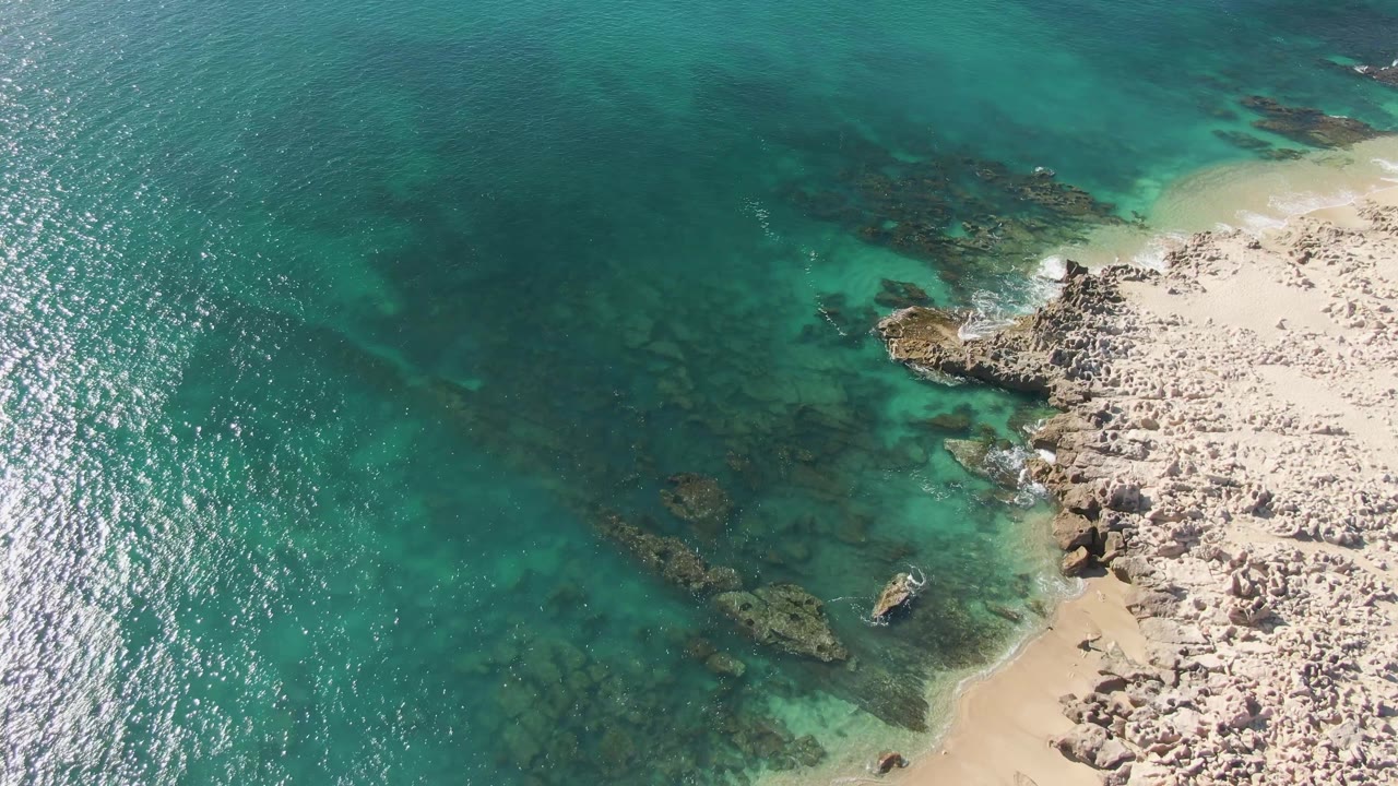 Aerial frame at Buzzards Beach showing green-blue shallow water around underwater rock formations, a narrow sandy curve, rocky coastal ground, bright water reflections, and no visible people.