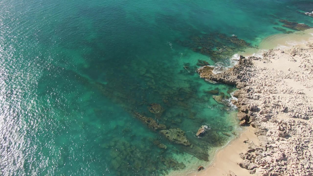Aerial frame over Buzzards Beach with darker submerged rock patches under turquoise water, a pale beach strip at the upper right, rocky shore at the right edge, small wave breaks, and no visible people.