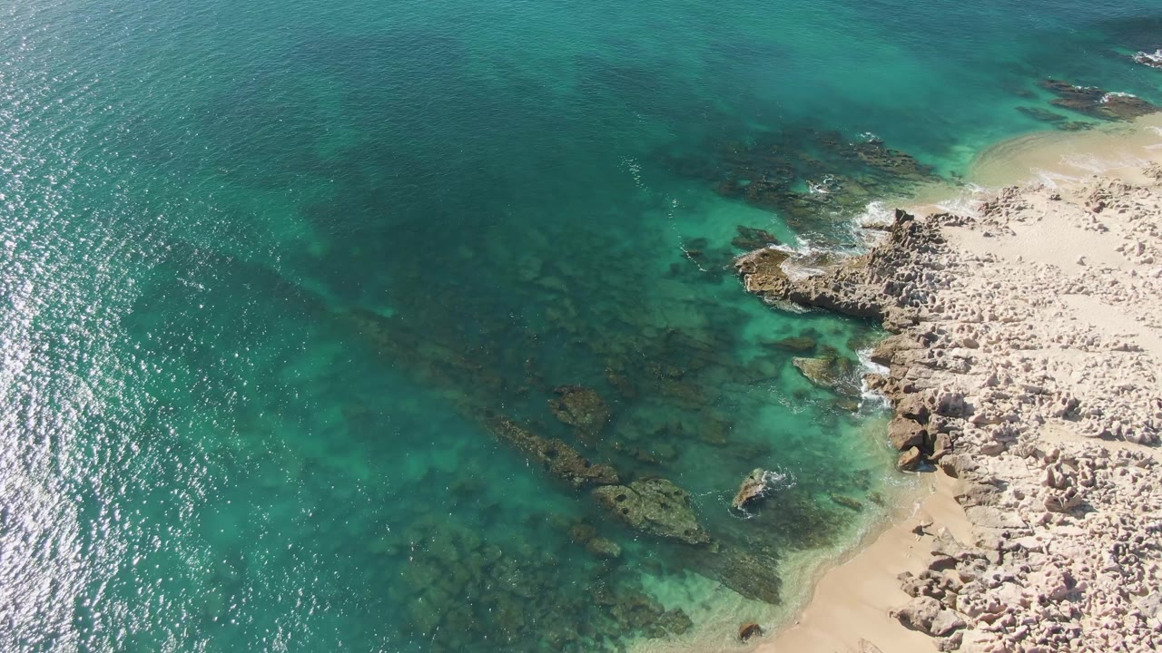 Aerial frame at Buzzards Beach showing a broader section of clear turquoise water, sunlit ripples, submerged rocks near center, sandy shoreline along the lower right, rocky coast, and no visible people.