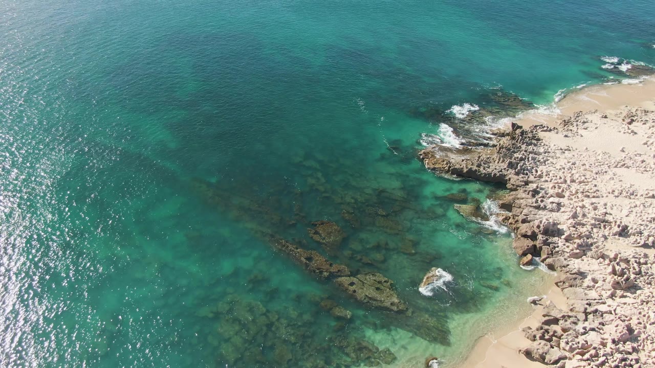 Aerial view of turquoise water, submerged rocks, pale sand, and foamy surf.