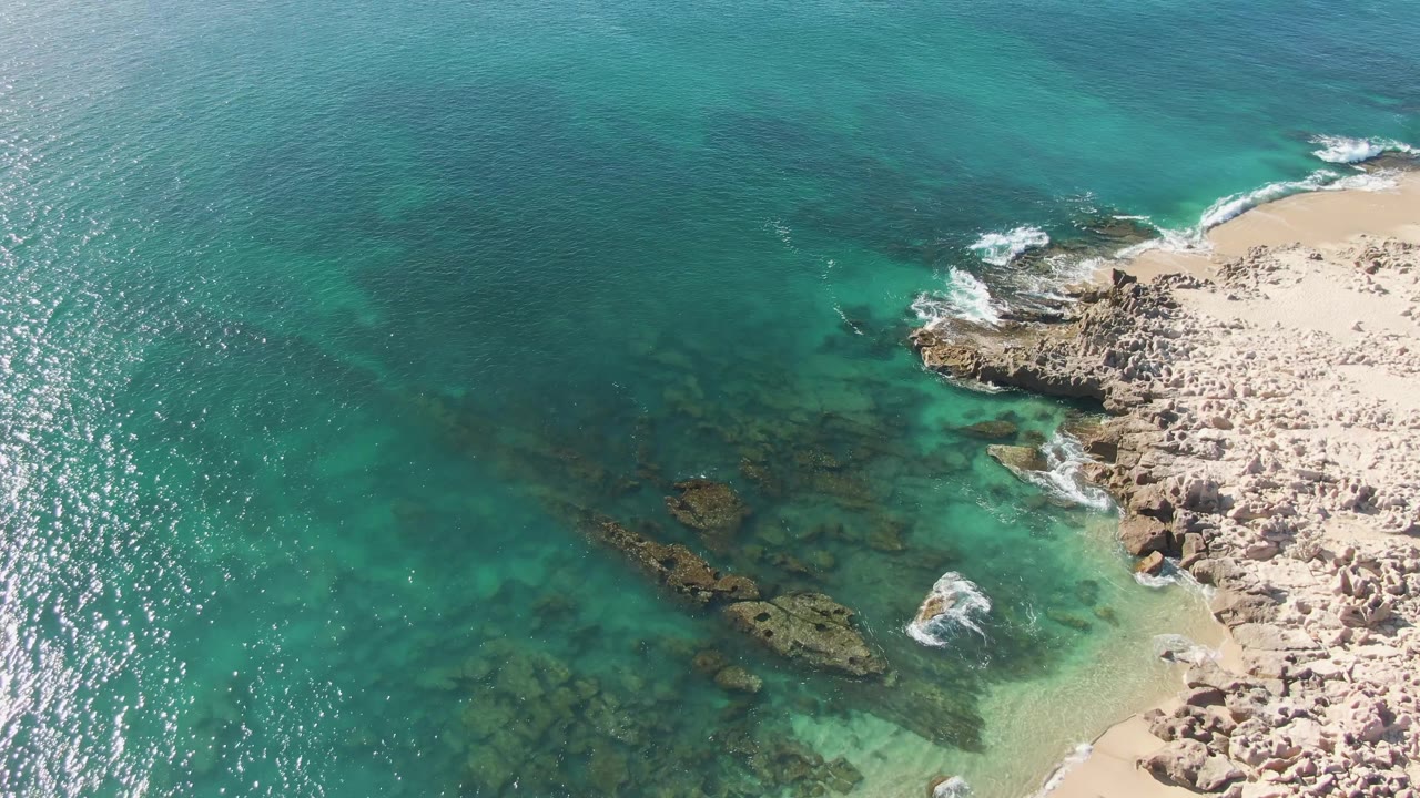 Aerial frame from Buzzards Beach, Baja California Sur, with bright sun glitter on turquoise ocean, dark submerged rocks, pale sand, foamy wave edges, and no visible people.