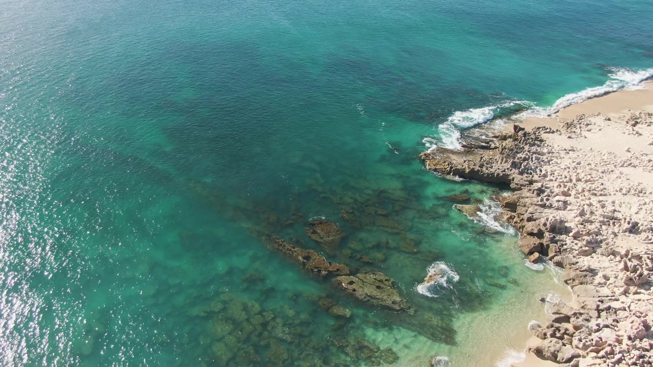 Aerial frame from Buzzards Beach, Baja California Sur, showing clear green-blue shallows, underwater rock patches, pale sand on the right, broken white surf, and no visible people.
