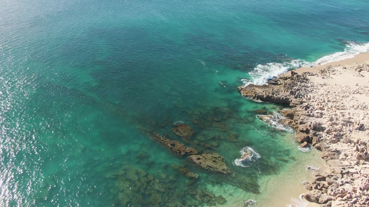 Aerial frame from Buzzards Beach, Baja California Sur, with turquoise water filling most of the view, rocks under the surface, a sandy rocky shoreline to the right, light foam, and no visible people.