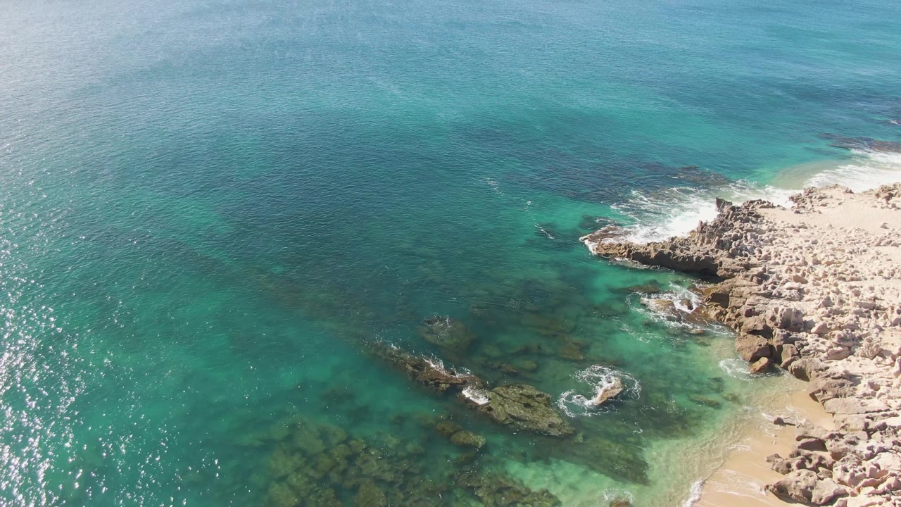 Aerial frame over Buzzards Beach in Baja California Sur showing turquoise shallows, darker submerged rocks, white foam on the shore, and no visible people.