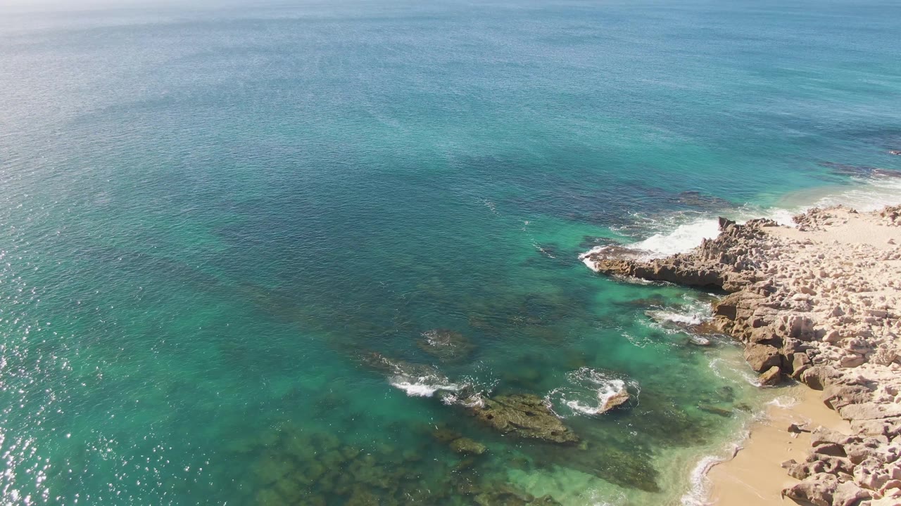 Aerial frame from Buzzards Beach showing deeper blue water across the top, turquoise shallows near submerged rocks, surf hitting the right shoreline, and no visible people.