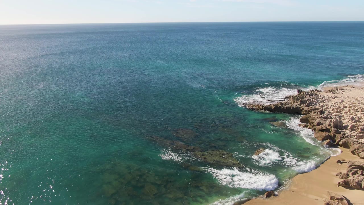 At 0:01, the Buzzards Beach shoreline remains on the right while white foam spreads farther across the rocks and sand below the turquoise shallows, with no visible people.