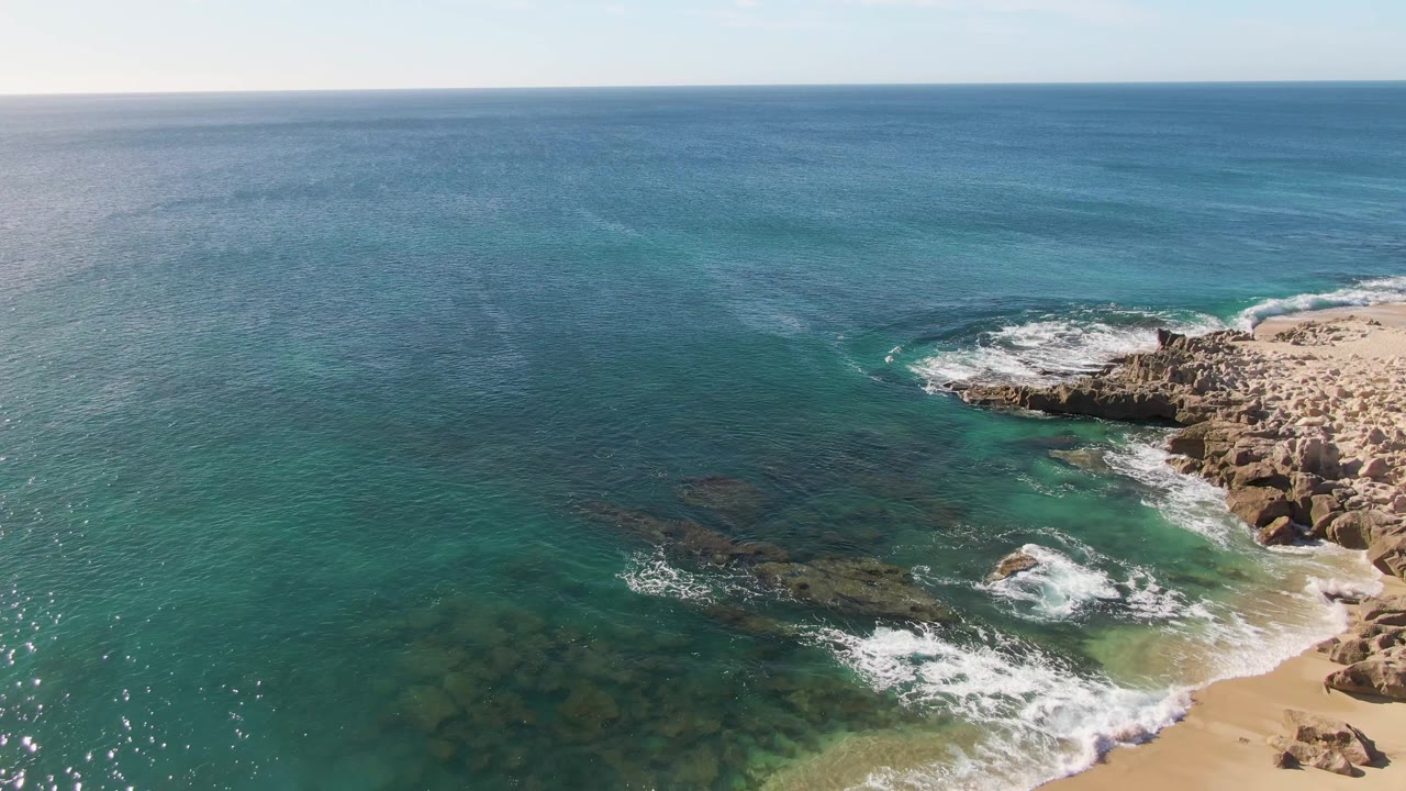 At 0:02, clear turquoise water exposes darker submerged rock patches while a fresh band of white surf curls around the rocky Buzzards Beach shore, with no visible people.