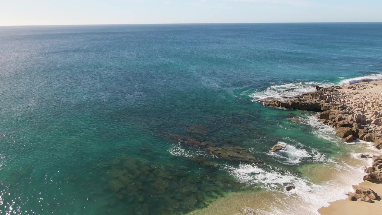 At 0:03, the sandy lower shoreline is more prominent, with white foam washing across the beach edge and rocky outcrops under bright water at Buzzards Beach, no visible people.