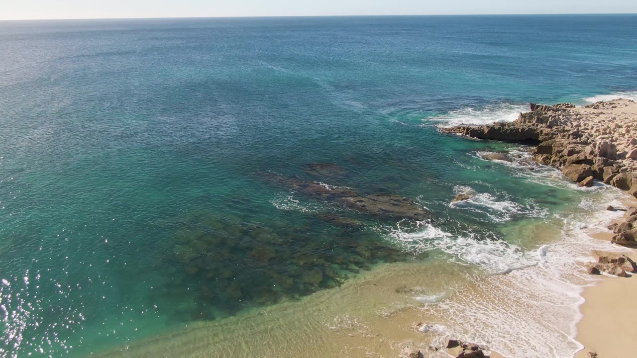 Aerial frame from Buzzards Beach, Baja California Sur showing turquoise shallows, darker offshore water, white foam near the sand, and rocky outcrops on the right with no visible people.