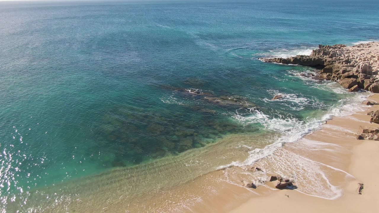 Buzzards Beach aerial frame with a wider band of exposed sand, white foam tracing the shore, darker underwater rock shapes offshore, and rocky outcrops at the upper right with no visible people.