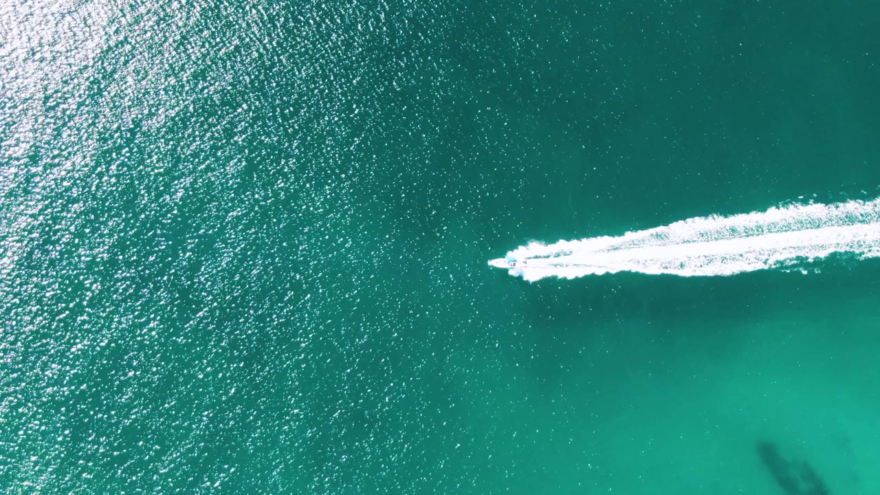 At Buzzards Beach, Baja California Sur, a small speedboat begins crossing turquoise water from left to right, leaving a white wake with no visible people.