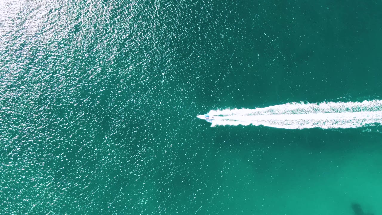 Near the right side of the frame, the boat continues over turquoise water at Buzzards Beach, with its white wake trailing left and no visible people.