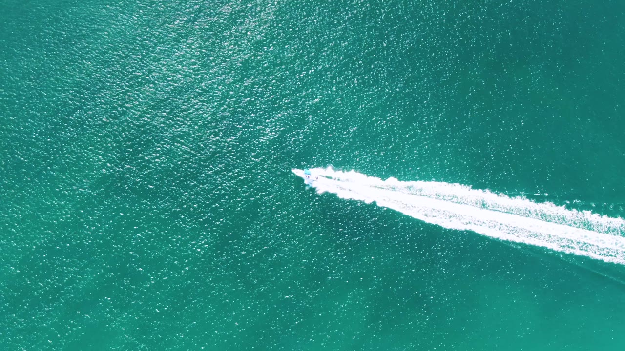 Overhead frame of the speedboat farther across the turquoise ocean at Buzzards Beach, Baja California Sur, Mexico, with its wake widening behind it and no visible people.
