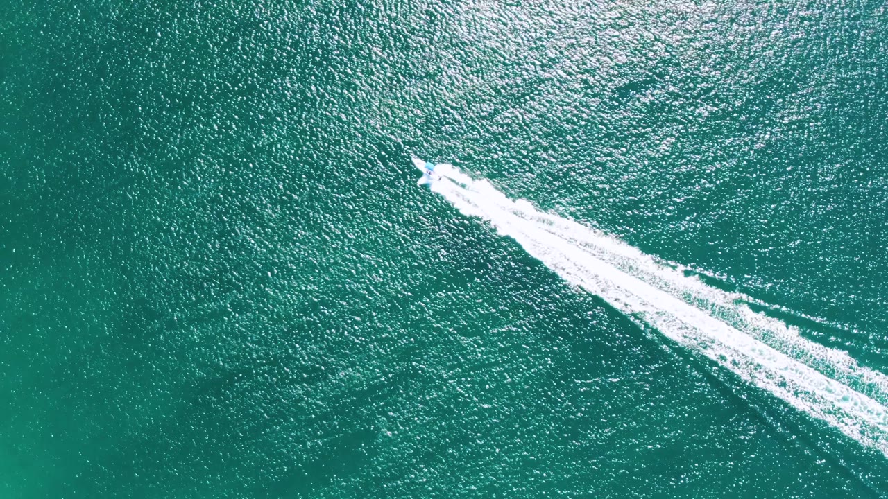 Aerial view at Buzzards Beach, Baja California Sur, Mexico, where the speedboat reaches the upper right of the frame and leaves a bright diagonal wake across textured water, with no visible people.