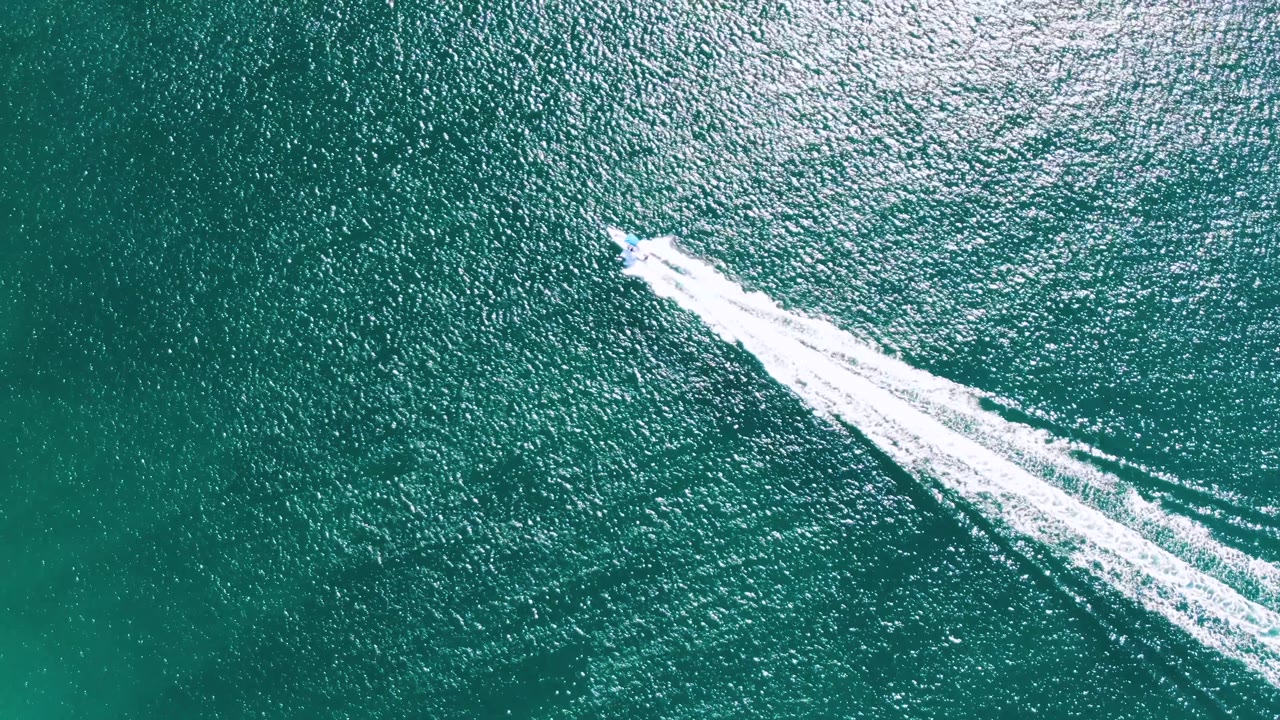 Overhead Buzzards Beach frame showing the speedboat continuing across turquoise ocean while its white foam wake trails diagonally through the center, with no visible people.