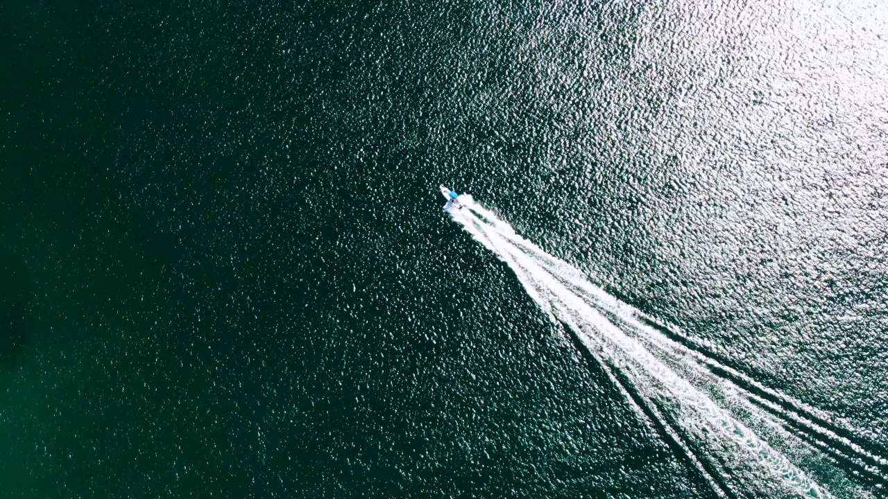Overhead shot of a small speedboat continuing across blue-green water near a brighter patch of reflected light in Buzzards Beach, with no visible people.