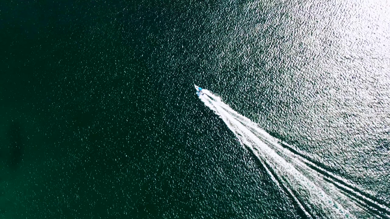 Aerial view at Buzzards Beach showing the speedboat advancing across darker blue-green ocean while its white wake fans out behind it, with no visible people.