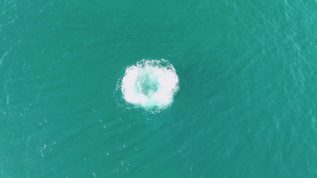 At Buzzards Beach in Baja California Sur, the splash widens into a brighter foamy circle on teal water near the center of the overhead frame, with no visible people.