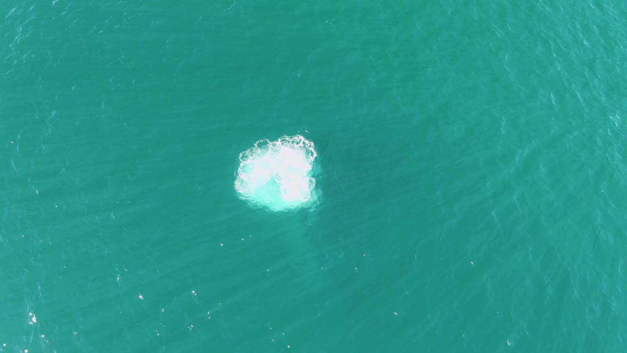 A larger white splash spreads across the turquoise ocean surface in the Buzzards Beach clip, with soft ripples radiating into otherwise clear water and no visible people.