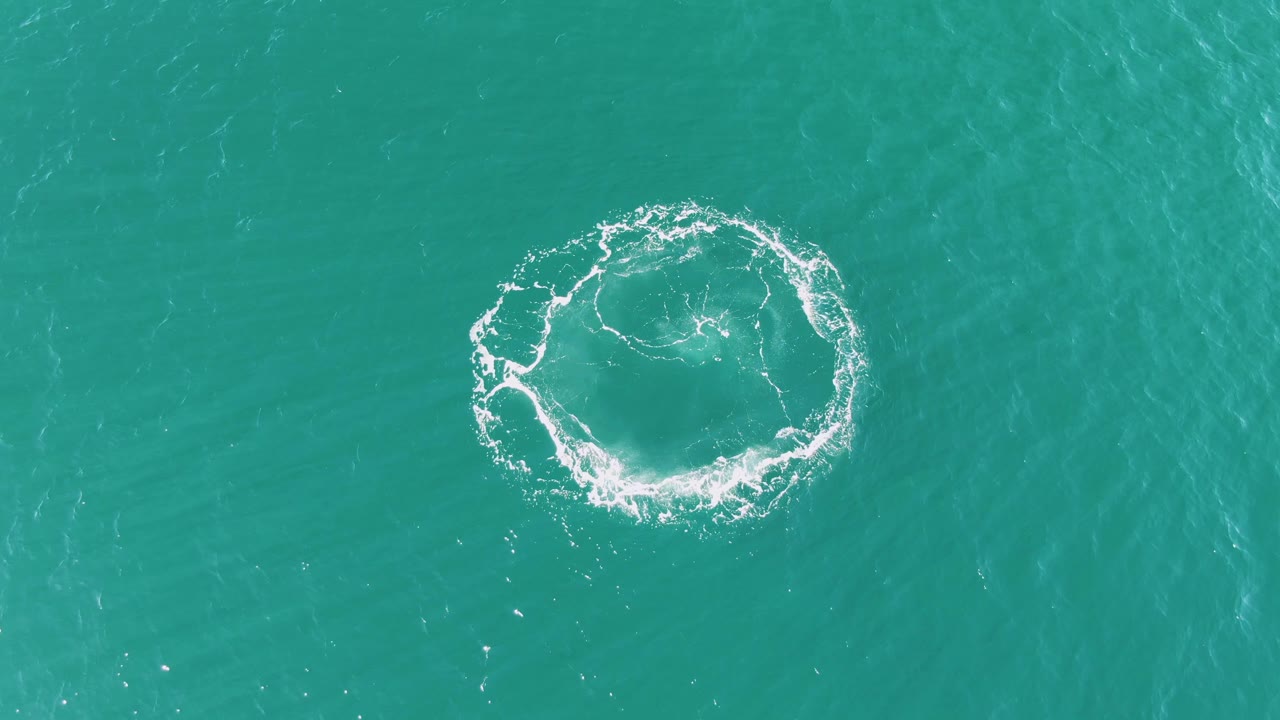 The foam circle remains centered over rippled turquoise ocean at Buzzards Beach, with a darker center and no visible people.