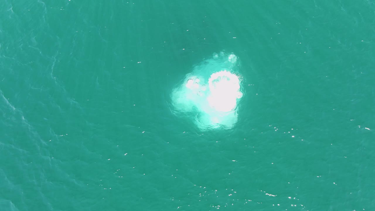 The white bubble patch is larger and brighter on the turquoise ocean surface at Buzzards Beach, with no visible people.