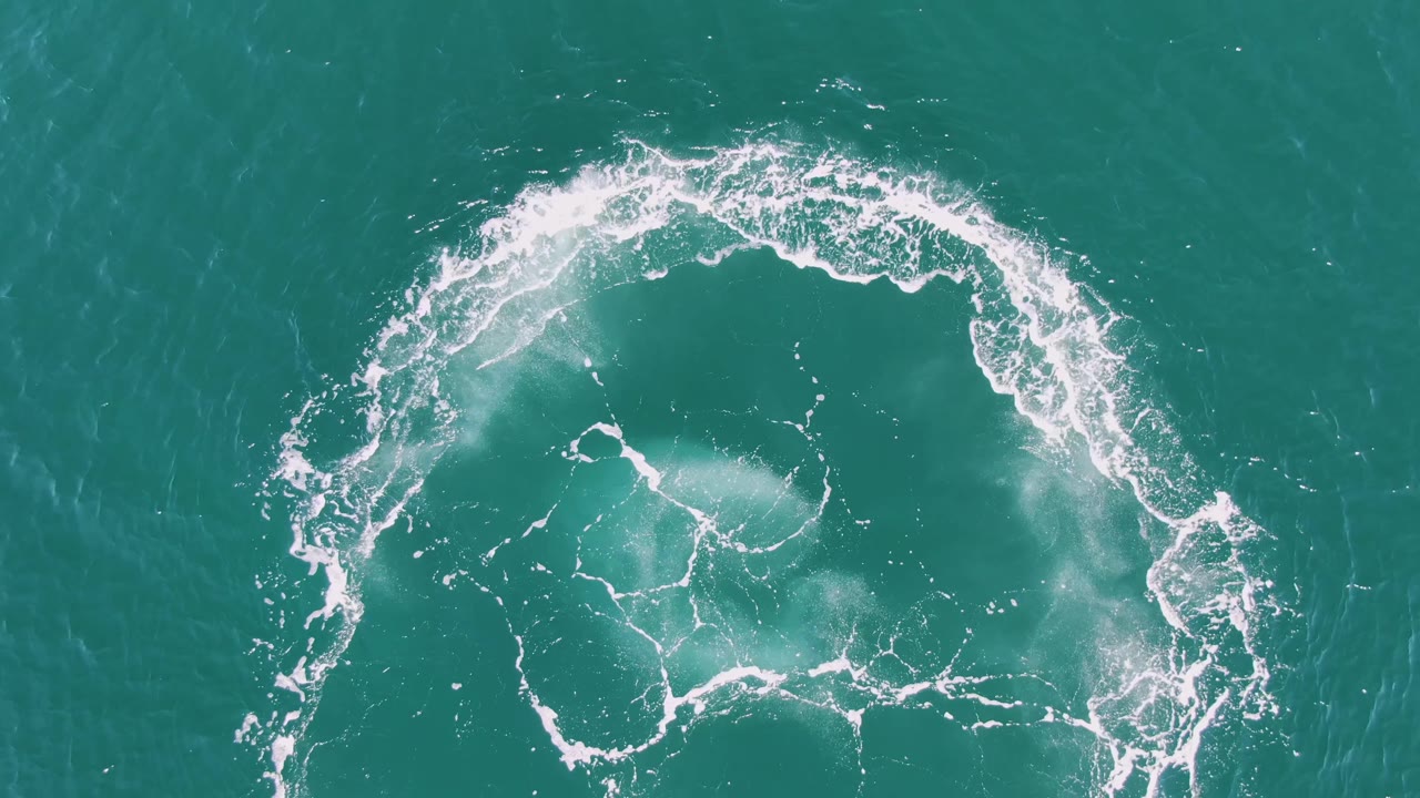 At two seconds, the Buzzards Beach aerial view shows the right side of the white foam ring breaking into smaller streaks over turquoise water, with no visible people.