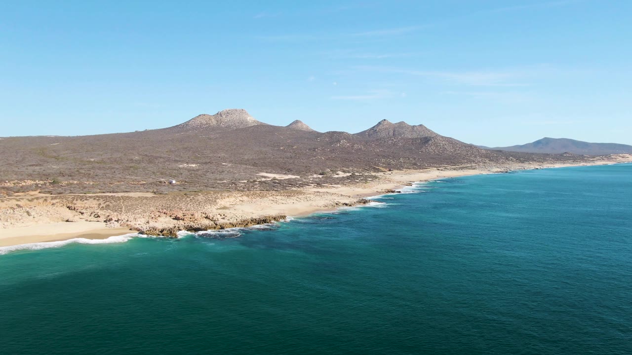 Buzzards Beach aerial view at 0:01 with deeper blue water in the foreground, a pale sandy shore, low rocks along the coast, distant mountains, and no visible people.