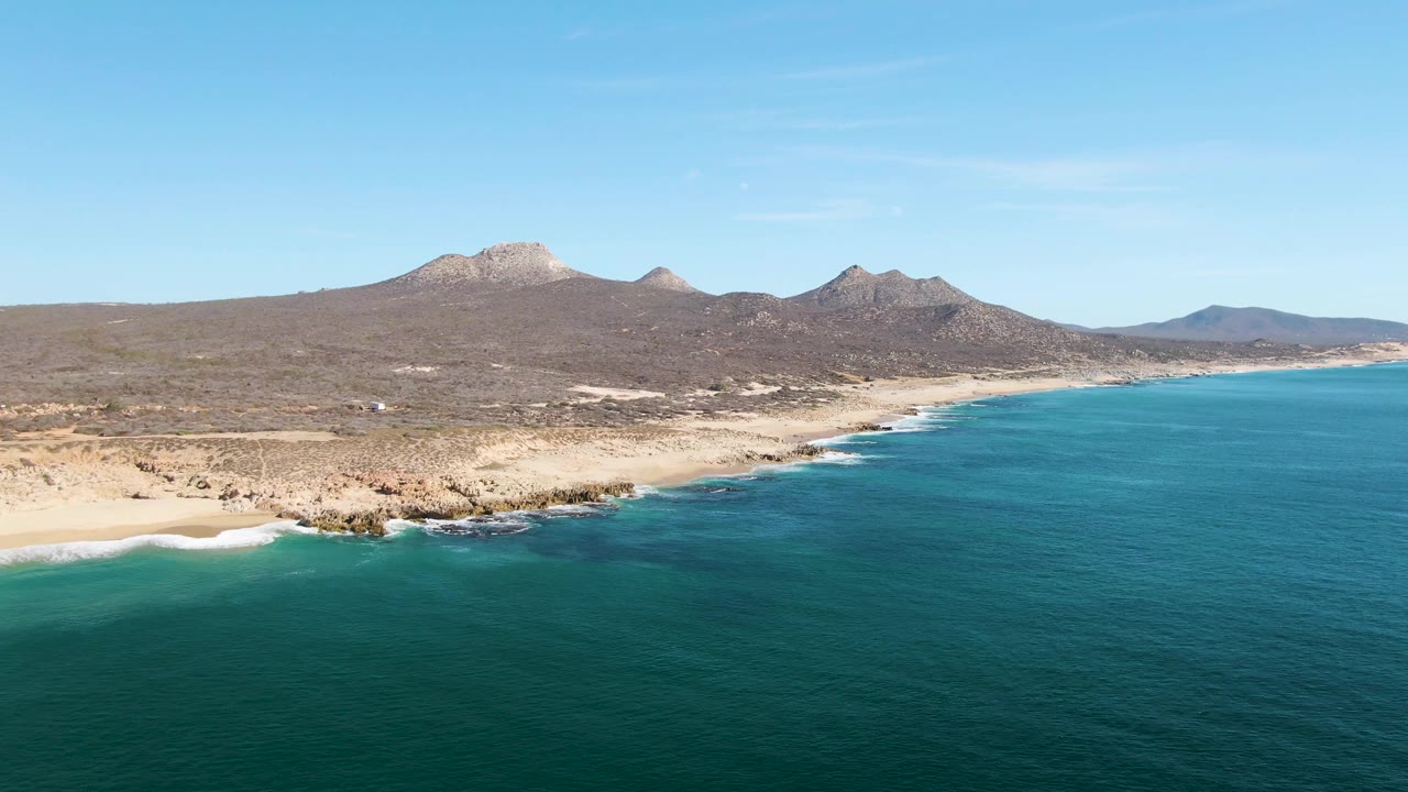 Buzzards Beach, Baja California Sur, seen from above with broad blue water, a narrow sandy beach, scattered shore rocks, brown coastal terrain, distant mountains, and no visible people.