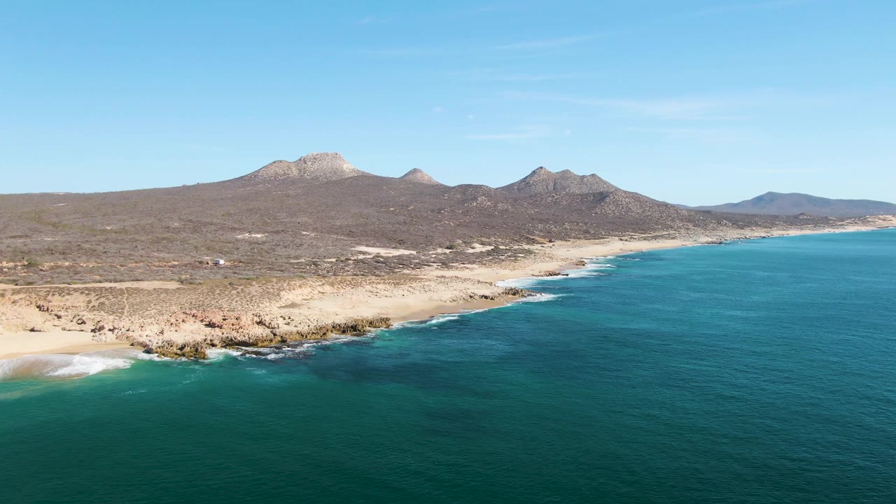 At Buzzards Beach in Baja California Sur, turquoise water fills the foreground beside a rocky sandy shoreline with dry mountains under clear sky, with no visible people.