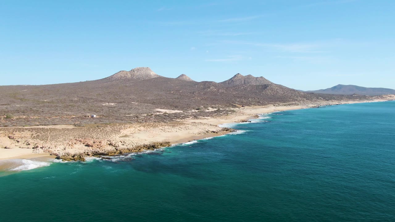 A second aerial frame holds the Buzzards Beach shoreline as white surf touches dark rocks and pale sand below distant Baja California Sur mountains, with no visible people.