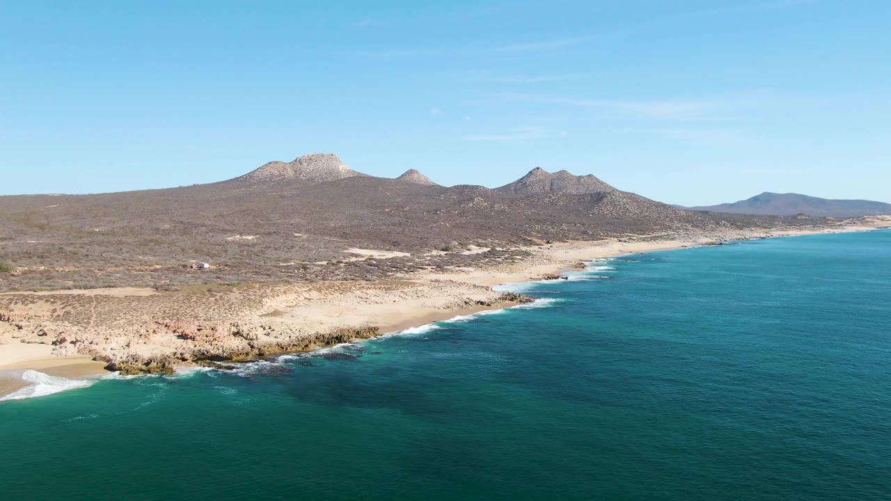 The Buzzards Beach coast remains in a broad aerial view, with deeper blue water to the right, rocky tan shore through the center, and low mountains across the horizon, with no visible people.