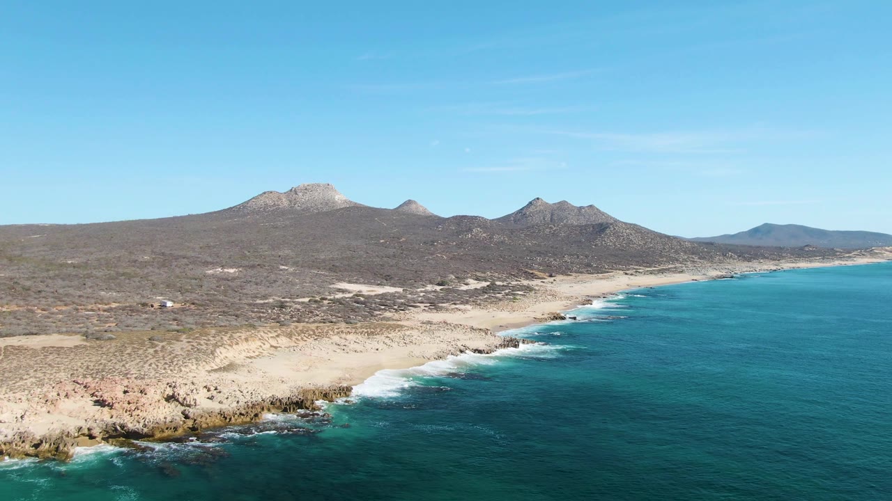 Aerial view over Buzzards Beach in Baja California Sur with turquoise ocean, white surf on a rocky sandy shoreline, dry scrubland, distant mountains, and no visible people.