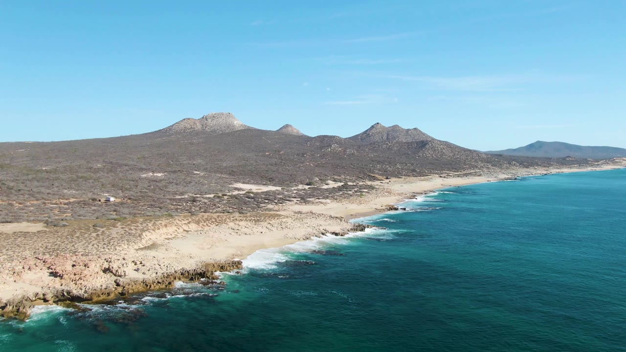 Wide aerial frame at Buzzards Beach showing blue-green water along the right edge, pale sand and rocks at the shore, brown scrubland inland, distant mountains, and no visible people.