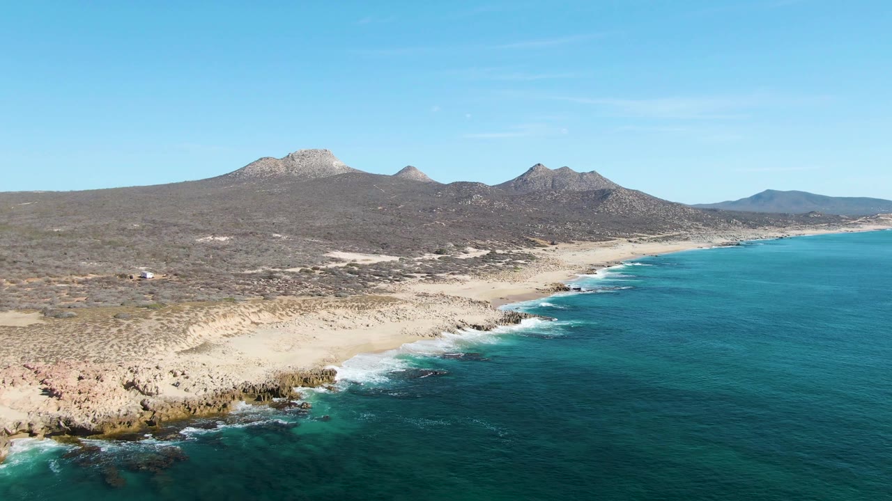 Aerial frame over Buzzards Beach with the shoreline running from lower left toward the distance, turquoise water to the right, desert terrain and mountains beyond, and no visible people.