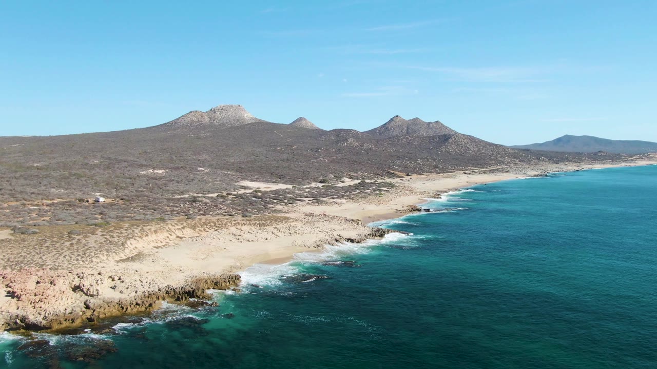 Drone view of rocky surf, sandy beach, scrubland, and mountains.