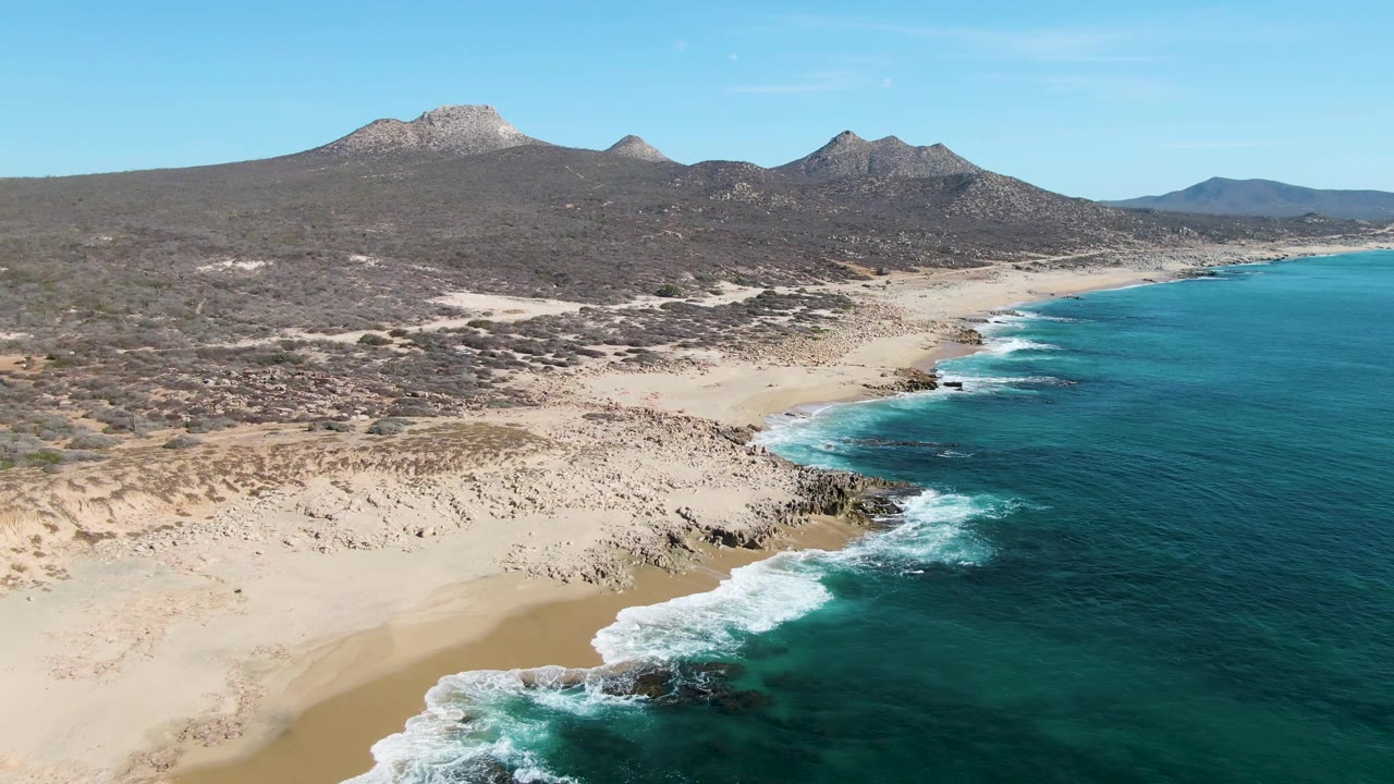 Aerial view of waves breaking along a rocky sandy coastline with mountains inland.