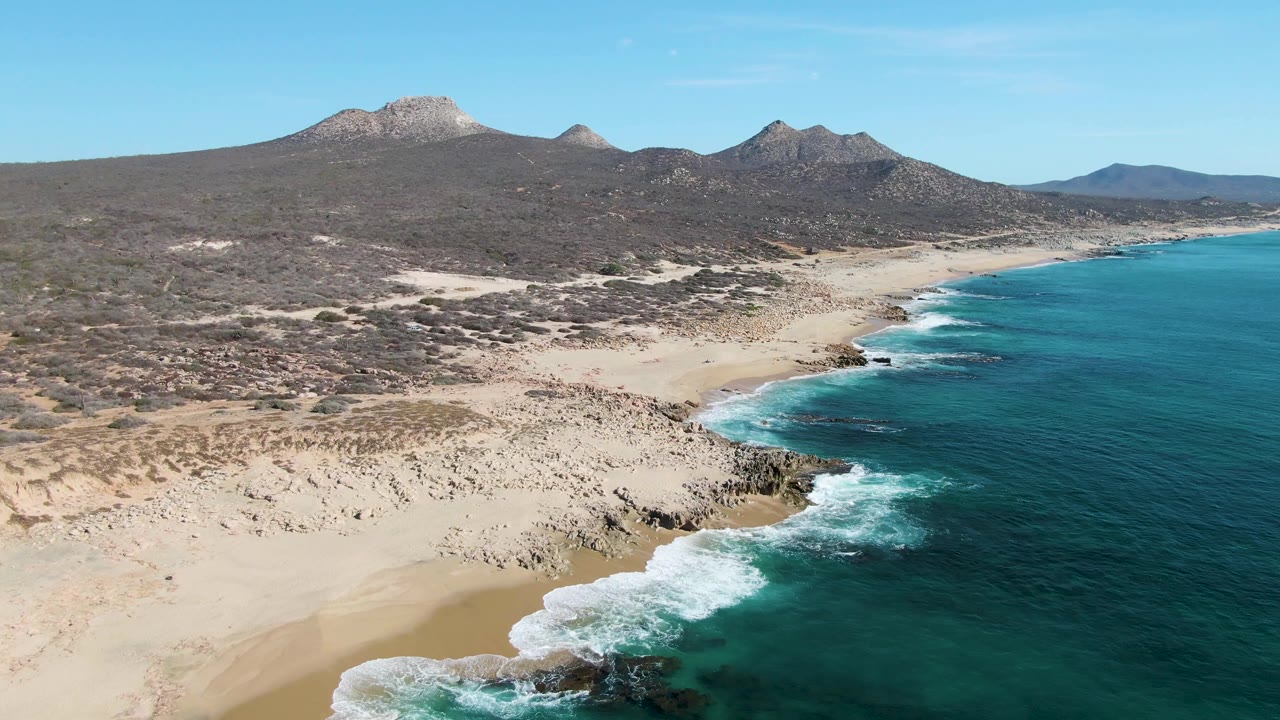 At 0:01, the aerial view holds over Buzzards Beach as a broader patch of white foam spreads along the rocks, with turquoise water to the right, pale sand below, mountain ridges inland, and no visible people.