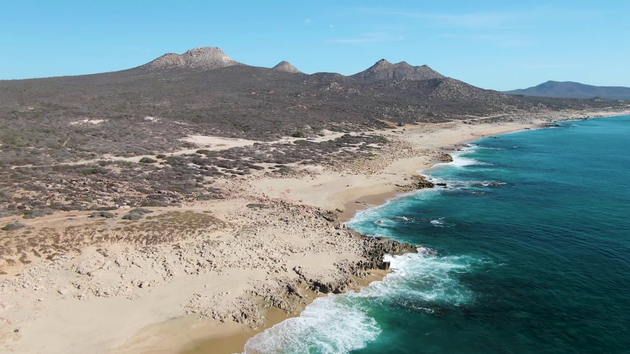 At 0:03, the aerial composition shifts slightly along Buzzards Beach, showing more open sand in the foreground, surf wrapping around dark shoreline rocks, scrub-covered terrain beyond, and no visible people.