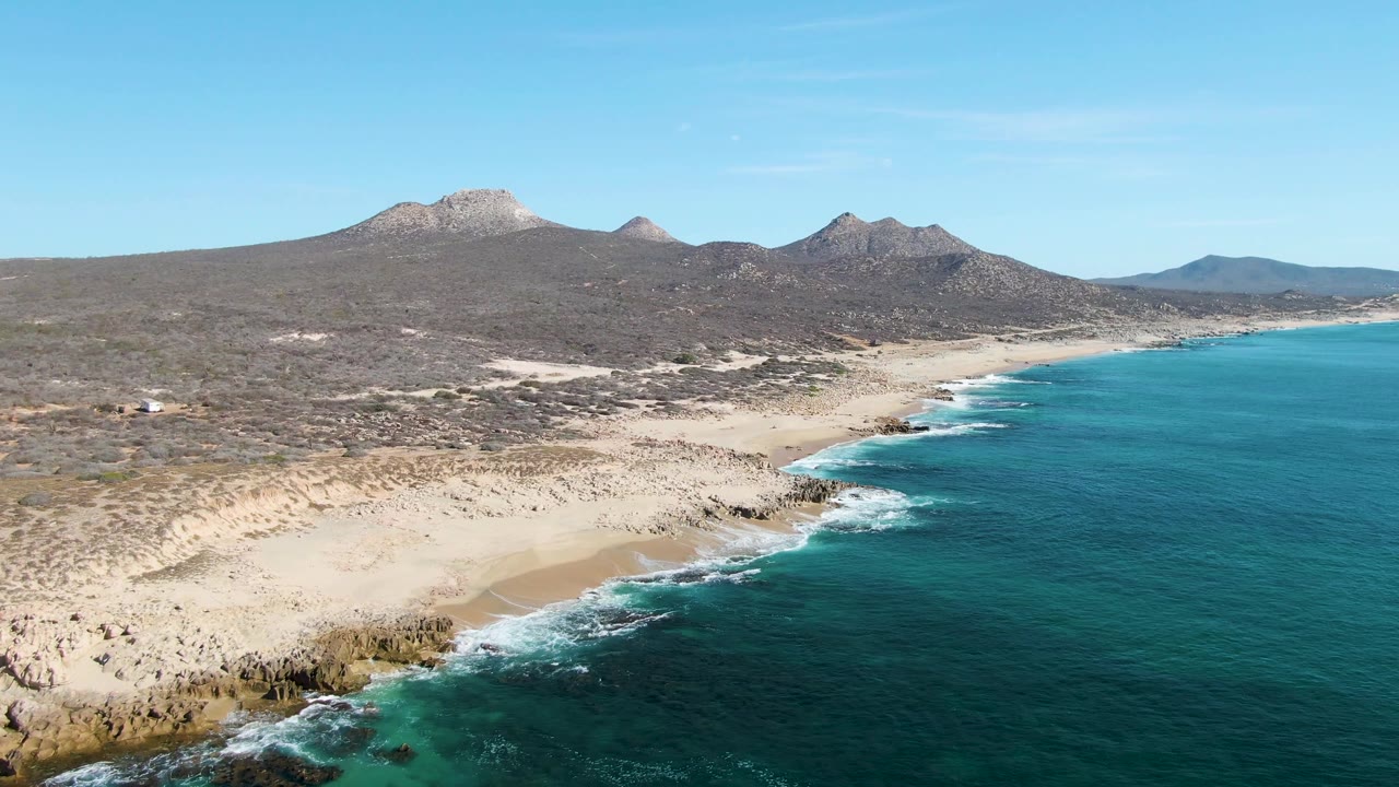 Aerial view over Buzzards Beach in Baja California Sur, Mexico, with turquoise ocean on the right, sandy beach and rocks below, arid mountains across the horizon, and no visible people.