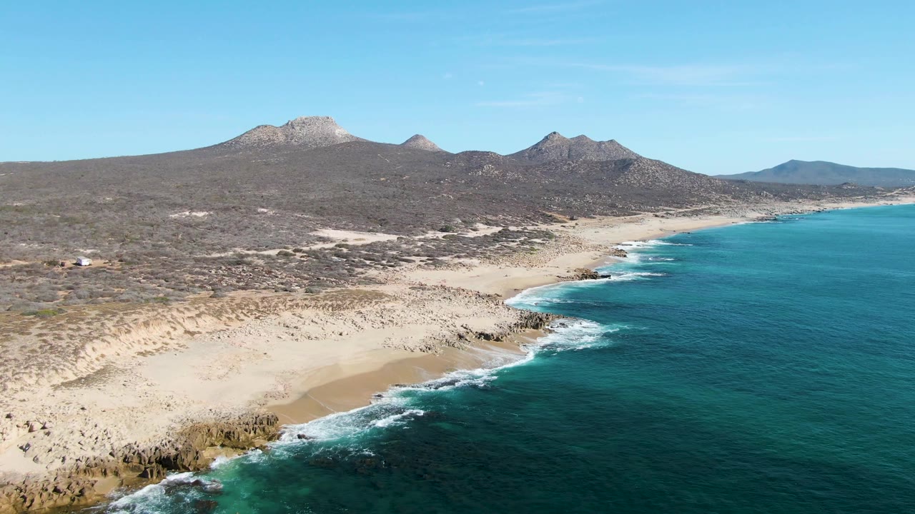 Wide aerial frame of Buzzards Beach showing white waves tracing the sandy shore, dark rocks near the lower coastline, dry scrubland behind the beach, and no visible people.