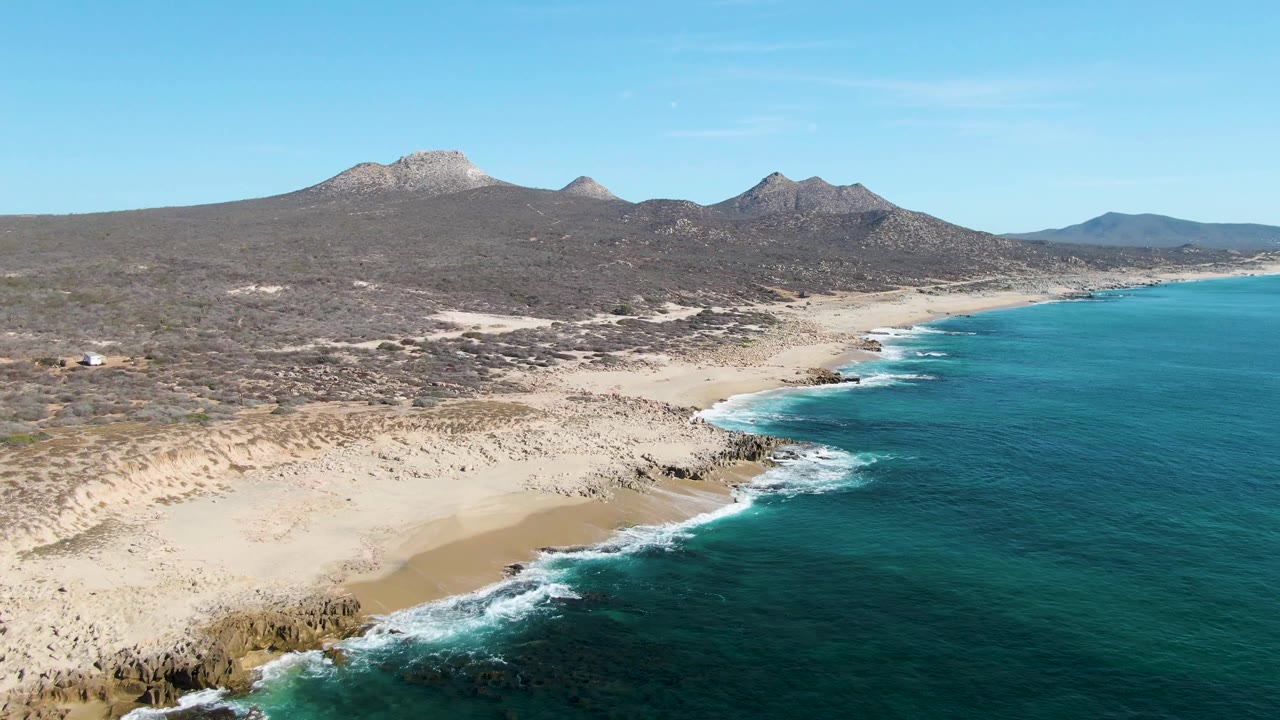 Aerial view at Buzzards Beach with the coastline curving away to the right, blue-green ocean filling the foreground side, pale sand and rocky points along the water, and no visible people.
