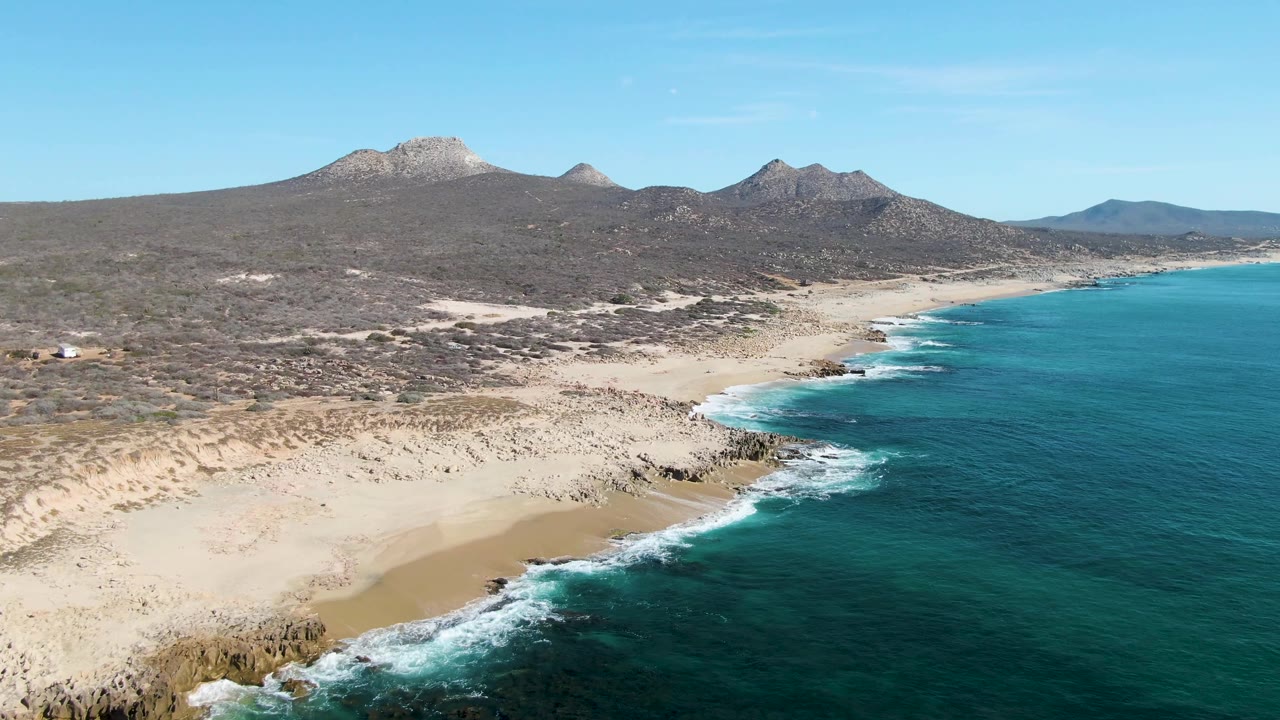 Aerial beach scene with turquoise water, rocks, desert scrub, and mountains